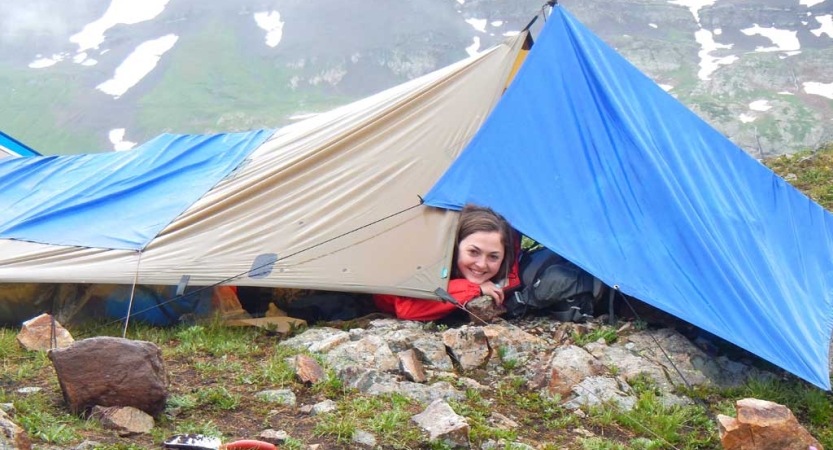 From under a tarp shelter, a person pokes their head out and smiles. Behind them, there appears to be a large mountain. 